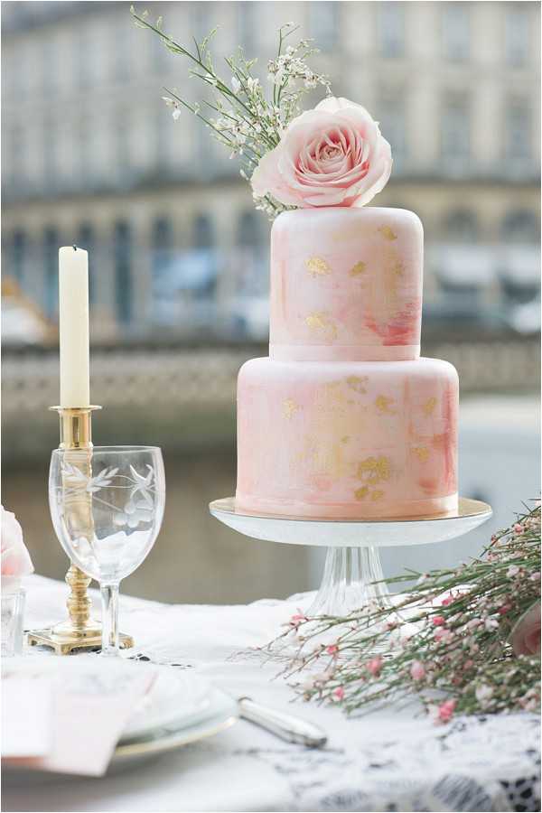 Close-up detail shot of a two-tiered wedding cake with a blush pink watercolor finish and gold leaf accents, topped with a single blush pink rose and sprigs of white waxflower. The cake is displayed on a white pedestal cake stand on a styled tablescape that includes an etched crystal wine glass, a brass taper candleholder with an ivory candle, pink waxflower stems, and white lace table linen. A blurred Haussmann-style building facade is visible in the background, suggesting an outdoor urban setting in Paris. The overall styling palette is blush pink and gold with a romantic, classic French aesthetic.
