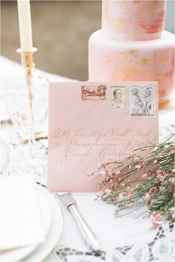 A close-up detail flat lay of a styled wedding tablescape featuring a blush pink envelope addressed in white calligraphy script to 'Mr and Mrs Paul West, 51 Pennsylvania Street, Denver, Colorado,' affixed with three vintage postage stamps. In the background, a two-tier blush pink and gold watercolor-painted wedding cake is partially visible, alongside a tall ivory taper candle in a gold candleholder, a cut crystal champagne flute, and a white plate with gold rim resting on white lace table linen. Sprigs of small pink wax flower blooms are scattered in the foreground near a silver cake knife. The overall decor palette is blush pink, ivory, and gold with a romantic classic styling theme.