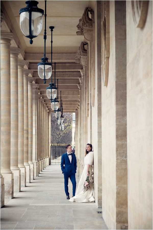 A couple portrait taken in a grand covered colonnade, likely the Palais Royal in Paris, featuring a long row of stone columns and a series of dark metal hanging lanterns that recede into the background. The groom walks along the colonnade wearing a navy blue suit with a white dress shirt and black bow tie, while the bride stands leaning against a column to the right wearing an ivory fitted gown with a white fur or feather-trimmed wrap and holding a bouquet with blush and muted tones. The composition is a wide portrait shot that uses the depth of the colonnade and repeating lanterns as strong leading lines, placing the couple small within the grand architectural setting. The styling is classic and polished, with a Parisian urban backdrop. Potential venue feature image.