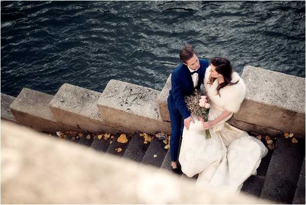 A couple portrait shot from an elevated overhead angle, showing the bride and groom seated together on stone quayside steps beside a dark river, likely the Seine in Paris. The groom wears a navy blue suit with a black bow tie, while the bride wears a white ball gown with a white fur stole wrap and holds a small bouquet of blush pink and dried lavender-toned flowers. The composition is a wide overhead shot capturing the stone steps, scattered autumn leaves, and the deep blue-grey water in the background, with the couple leaning close together looking downward.