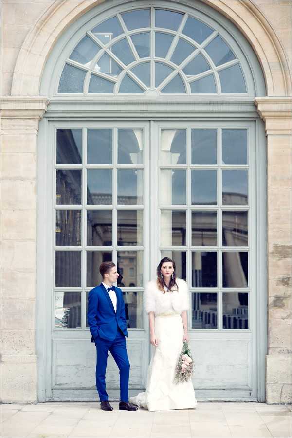 A couple poses for a portrait in front of a large arched pale blue-grey double door with a fanlight window, characteristic of classic Parisian architecture. The groom wears a cobalt blue suit with a white dress shirt and black bow tie, while the bride wears a fitted ivory gown with a white faux fur or feathered capelet and holds a loose, trailing bouquet of blush pink and pale blooms. The two stand slightly apart, facing each other, against the ornate stone facade. The shot is a full-length portrait composition with the architectural doorway as a prominent backdrop. Potential venue feature image.