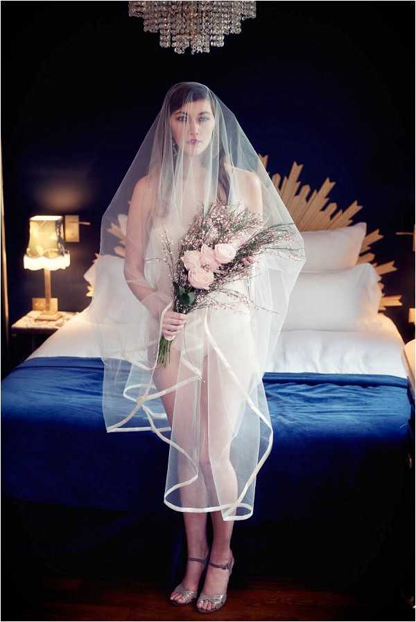A bridal boudoir-style portrait of a bride standing in a darkly decorated hotel or bedroom interior. She is wearing a sheer, floor-length veil with a satin ribbon edge that covers her body, silver strappy heeled sandals, and holds a loose bouquet of blush pink roses and wispy dried or fine-stemmed filler flowers. The room features deep navy blue walls, a crystal chandelier overhead, a gold sunburst headboard on the bed with white bedding and a royal blue base, and a warm-toned table lamp on the nightstand. The composition is a full-length portrait shot with dramatic, moody lighting creating strong contrast between the dark background and the white veil.