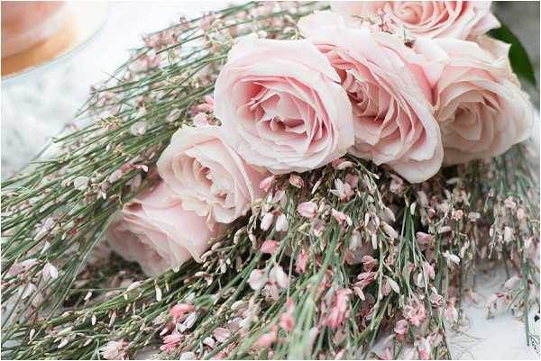Close-up detail shot of a bridal bouquet or floral arrangement featuring soft blush pink garden roses paired with cascading sprigs of pale pink limonium (statice) and fine green stems. The flowers are laid on a light surface, with a blurred white object visible in the upper left corner suggesting a cake or glassware nearby. The overall palette is soft blush and pale pink with green accents, consistent with a romantic, classic wedding styling aesthetic.