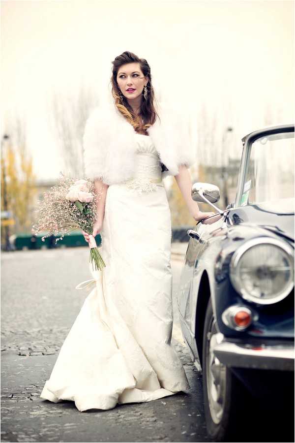 A bridal portrait taken outdoors on a cobblestone street, featuring a single bride standing beside a vintage dark navy convertible car. The bride wears a fitted ivory satin mermaid-style wedding gown with a beaded or embellished waistband, paired with a white fur stole wrap. She holds a loose bouquet of blush pink roses and baby's breath tied with a light pink ribbon. Her dark hair is styled in a soft updo, and she wears statement drop earrings with bold red-pink lipstick, giving a retro 1950s–1960s styling influence. The image is shot as a medium-full portrait with a shallow depth of field, with the background softly blurred. The overall color treatment has a faded, warm-toned vintage quality.