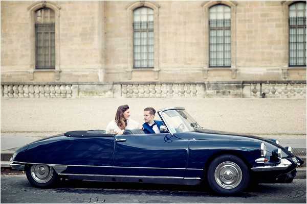 A couple poses in a navy blue vintage Citroën DS convertible parked on a Parisian street, with a large classical stone building featuring tall arched windows and a balustrade visible in the background. The bride wears a white dress and the groom wears a navy suit with a blue bow tie, and they are turned toward each other in conversation. The shot is a wide portrait taken from street level, capturing the full length of the classic French car. The overall styling is classic and urban, with the vintage vehicle serving as a deliberate design element complementing the Parisian architectural backdrop.