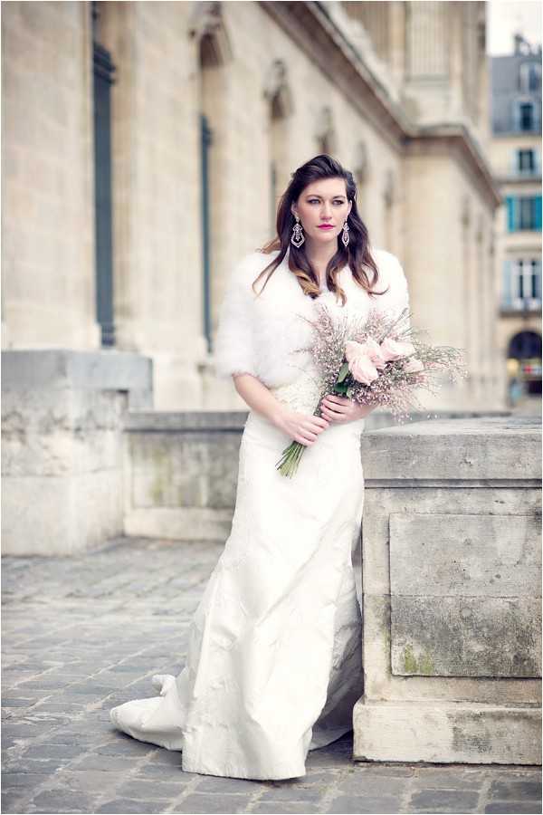 A bridal portrait taken outdoors in a Parisian urban setting, with classic Haussmann-style architecture and cobblestone pavement visible in the background. The bride stands alone wearing a fitted ivory gown with a train and a white faux-fur shrug wrap, accessorized with chandelier earrings and bold pink-red lipstick. She holds a loosely gathered bouquet of blush pink roses and dried pink limonium with exposed green stems. The image is a medium full-length portrait with a shallow depth of field that softly blurs the architectural backdrop.