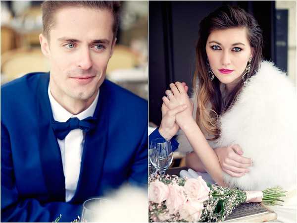A split-image portrait of a couple, likely at a reception table. On the left, a close-up of the groom wearing a navy blue tuxedo jacket with a black bow tie and a small floral boutonniere, looking slightly off-camera. On the right, the bride is seated at the table wearing a white feather-trimmed wrap or stole, with bold pink lipstick, dangling gold earrings, and loosely styled ombre hair; she holds the groom's hand and gazes directly at the camera. A bouquet of blush pink roses with greenery and a wine glass are visible on the table in the foreground. The overall styling is classic with a modern edge, featuring a cool blue and blush pink color palette.