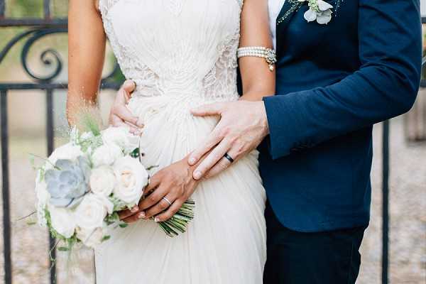 A close-up portrait of a bride and groom standing together, cropped from the neck to the hips. The bride wears an ivory beaded and lace gown with a flowing skirt, a pearl bracelet, and holds a bouquet of white garden roses, dusty blue-green succulents, and soft greenery with wispy grasses. The groom wears a navy blue suit jacket with dark trousers and a black wedding band, and has a small white floral boutonniere with greenery. His hands are placed around the bride's waist. Both wedding rings are visible. The setting appears to be outdoors near a wrought iron gate, with a classic or rustic styling approach.
