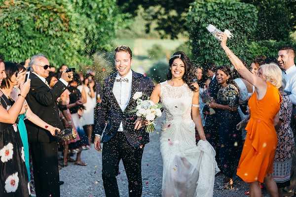 A newlywed couple walks through a confetti exit along a tree-lined gravel path, surrounded by approximately 20-30 guests on either side cheering and throwing colorful confetti. The groom wears a navy suit with a floral bow tie, while the bride wears a lace-detailed ivory gown and carries a white and cream bouquet, with a small hair accessory. The outdoor setting features a formal avenue of tall, trimmed trees in warm late-afternoon sunlight. The composition is a wide portrait shot taken straight down the path, capturing the couple mid-stride and laughing as confetti fills the air around them.