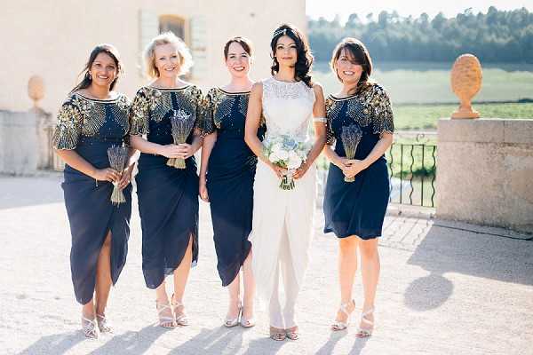 A bridal party portrait taken outdoors on a stone terrace in front of a French chateau, featuring the bride and four bridesmaids. The bride wears a white sleeveless two-piece outfit with a lace crop top and wide-leg trousers, accessorized with a delicate headpiece, and holds a bouquet of white and pale blue flowers including what appear to be white roses and dusty blue thistles or hydrangeas. The four bridesmaids wear matching navy midi dresses with gold and navy beaded, embellished cap sleeves, paired with silver strappy heels, and each holds a small bundle of dried lavender tied with twine. The styling has a classic Art Deco-inspired feel, with the beaded embellishments on the bridesmaids' dresses adding gold and silver detail to the navy palette. The image is a wide portrait shot taken in bright natural sunlight.
