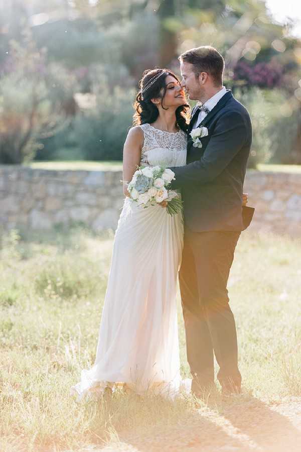 A couple portrait taken outdoors in a sun-drenched garden setting, with the groom leaning in to kiss the bride on the forehead or temple. The bride wears a floor-length ivory gown with a lace illusion neckline and flowing chiffon skirt, accessorized with a delicate beaded headpiece and her dark hair styled in loose waves. She holds a rounded bouquet of blush roses, white roses, and green succulents with palm leaf accents. The groom wears a dark navy suit with a bow tie and a white boutonniere. The composition is a full-length portrait with warm golden backlight creating a soft lens flare, suggesting late afternoon sun. The styling leans toward a romantic, slightly bohemian aesthetic with the embellished headpiece and succulent bouquet details.