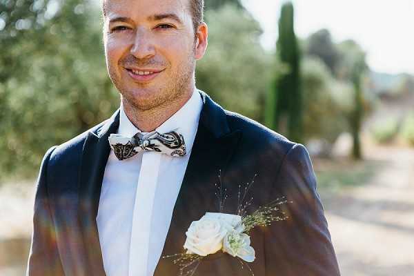 A close-up portrait of a groom outdoors, smiling directly at the camera. He is wearing a navy blazer with a white dress shirt and a black-and-white paisley-patterned bow tie, with a boutonniere featuring a cream rose and delicate dried grass sprigs pinned to his lapel. The background shows a sun-drenched outdoor setting with cypress trees and olive trees softly blurred. The shot is a mid-crop portrait with warm natural lighting and slight lens flare visible.