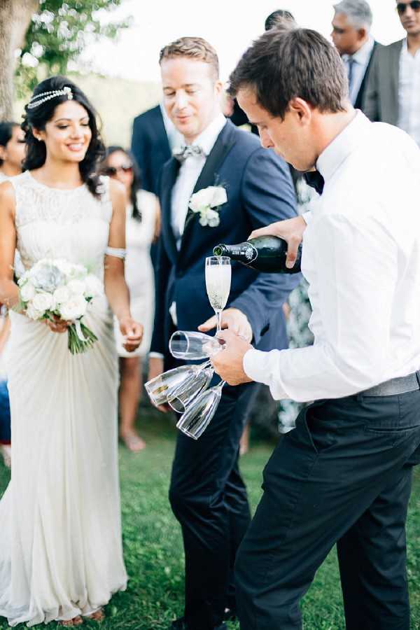 An outdoor cocktail hour or post-ceremony moment captured in a medium portrait shot, showing a waiter in a white shirt and black trousers pouring champagne from a dark bottle into flutes while balancing several glasses in one hand. The bride stands to the left wearing a draped, embellished ivory gown with a jeweled headband and holding a bouquet of white roses and greenery, smiling warmly. The groom stands beside her in a navy suit with a patterned bow tie and a white boutonniere. Several guests are visible in the background, and the setting appears to be an outdoor garden or lawn with trees, photographed in bright natural daylight.