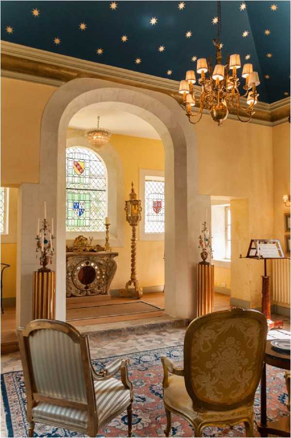 Interior shot of a historic chapel or oratory, likely within a French chateau, showing no people or wedding activity but capturing the architectural and decorative features of the space. The foreground room features a deep navy ceiling painted with gold stars, a brass chandelier with cream lampshades, warm yellow-ochre walls, two Louis XV-style armchairs upholstered in striped grey and gold damask fabric, and a patterned red and blue Persian-style rug. Through a stone rounded arch, a smaller chapel sanctuary is visible with heraldic stained glass windows in blue, green, red, and gold, an ornate carved altar, tall gilded candlesticks, and a crystal chandelier. The composition is a wide interior shot taken from a standing height. Potential venue feature image.