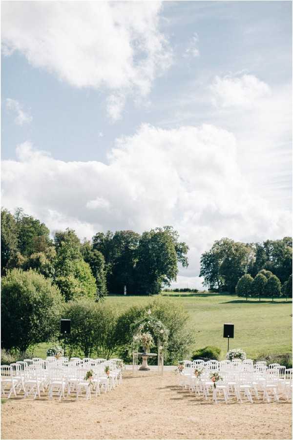 An outdoor wedding ceremony setup photographed before guests arrive, captured in a wide shot showing the full layout. White Napoléon chairs are arranged in two sections with a central aisle on a gravel or sandy surface, with small floral arrangements in blush and coral tones decorating the aisle chairs. At the front, a floral arch or altar structure is visible, adorned with flowers in soft pink and white tones, alongside what appears to be a stone pedestal or fountain. The setting opens onto a broad green lawn with mature trees in the background, giving the space a classic French country estate feel. Two black speaker stands are positioned on either side of the ceremony space. Potential venue feature image.