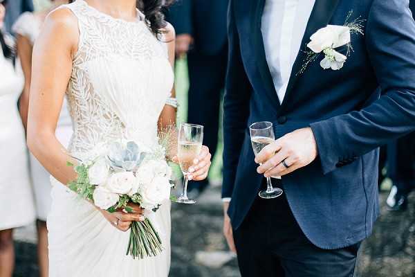 A close-up portrait of a bride and groom during what appears to be a cocktail hour or post-ceremony reception, each holding a champagne flute. The bride wears a fitted sleeveless ivory gown with intricate beaded lace detailing and holds a loose, garden-style bouquet of white roses, succulents, and eucalyptus foliage. The groom wears a navy blue suit jacket with a white shirt and a boutonniere of white flowers with greenery accents, and a dark wedding band is visible on his hand. Guests are softly visible in the blurred background, suggesting an outdoor setting with bright natural light.