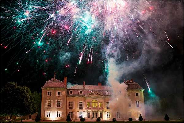 A fireworks display erupts above a large French château during a wedding celebration, photographed at night. The château is a symmetrical, multi-story stone manor with lit windows, ornamental topiary, and classic French architectural details including dormer windows and decorative cornices. Bursts of teal, green, pink, red, and white fireworks fill the dark sky above the building, with smoke billowing from launchers positioned at ground level in front of the façade. Wide-angle shot capturing the full building and the fireworks display in their entirety. Potential venue feature image.