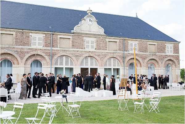 A cocktail hour is taking place outdoors on the terrace and lawn of a large French brick building with arched windows, a slate roof, and a central clock pediment. Approximately 40-50 guests dressed in formal attire — primarily dark suits and floor-length gowns in neutral and dark tones — are mingling in groups along the terrace. White folding chairs are scattered across the lawn in the foreground, along with what appears to be a wooden easel displaying a seating chart or welcome sign. The overall decor palette is white and neutral, with white furniture and floral arrangements visible on the terrace. Wide shot taken from the lawn. Potential venue feature image.