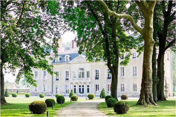 Wide exterior shot of a French chateau viewed from the grounds, with a gravel pathway leading directly to the main entrance. The building is a classic French manor house with light stone facade, symmetrical shuttered windows, a mansard roof with dormer windows, and a decorative wrought-iron balcony above the front door. The formal grounds feature manicured spherical topiary bushes lining the pathway and a lawn bordered by mature trees. No people are visible in the frame. Potential venue feature image.