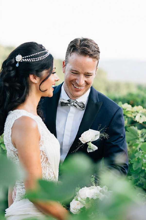 A couple portrait taken outdoors in what appears to be a vineyard setting, with lush green foliage framing the foreground. The bride wears a sleeveless lace-detailed white gown and an ornate pearl and crystal headpiece draped across her dark upswept hair, while holding a bouquet of pale blush and ivory roses. The groom is dressed in a navy tuxedo jacket with a white dress shirt and a patterned grey bow tie, and wears a white rose boutonniere with greenery. Both are laughing and facing each other in a candid, relaxed moment captured in a medium portrait crop with soft natural light.