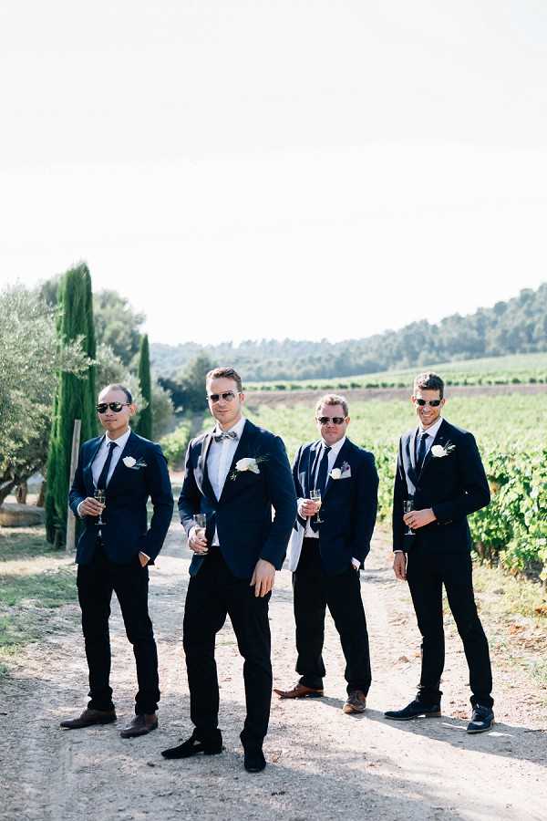 A groomsmen portrait taken outdoors on a gravel path at a vineyard, with rows of grapevines and cypress trees visible in the background. Four men are posed together — the groom is distinguished by a grey bow tie and white boutonniere, while the three groomsmen wear navy suits with dark ties and matching white boutonnieres. All four wear sunglasses and hold champagne flutes. The styling is modern and classic, with a consistent navy suit palette across the group. Medium full-length group shot.
