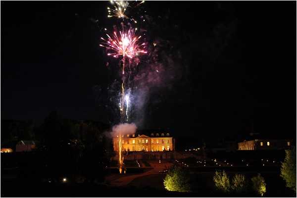 A wide-angle nighttime shot capturing a fireworks display above a large illuminated French chateau or manor house. The building is warmly lit with golden exterior lighting, and a trail of small lights lines the driveway or pathway leading up to the entrance. A single firework has burst in pink and purple tones high above the roofline, with smoke visible at the launch point directly above the building. The surrounding grounds are dark, with tree canopies faintly visible in the foreground. Potential venue feature image.