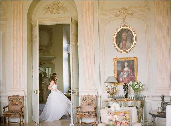 A bride in a white strapless ballgown with a full tulle skirt stands in a doorway, appearing to adjust or move her dress, captured in a wide interior portrait shot. The setting is a formal French chateau salon featuring ornate white and gold boiserie wall paneling, arched doorways with decorative carved moldings, and two 18th-century-style oil portrait paintings in gilded frames mounted on the wall. The room is furnished with gilt-framed armchairs upholstered in floral tapestry fabric, a console table with a lamp, and a small arrangement of white and blush flowers. The overall decor palette is cream, gold, and soft sage, consistent with a classic French chateau interior. Potential venue feature image.