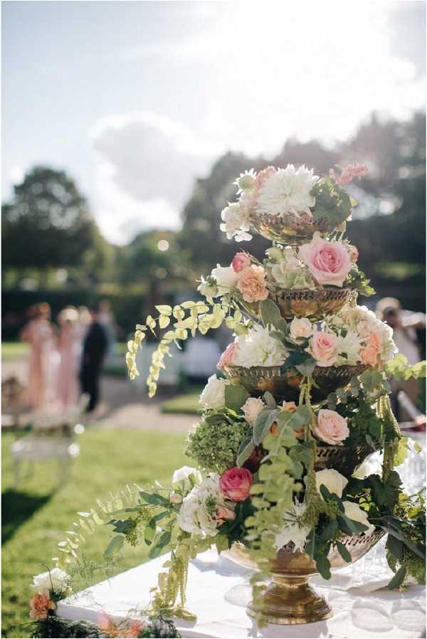 Close-up detail shot of a tiered brass urn floral arrangement displayed on a white-clothed table at an outdoor garden wedding. The three-tiered antique gold stand is densely arranged with blush and deep pink roses, peach carnations, white hydrangeas, cream dahlias, green viburnum, and trailing eucalyptus and fern foliage. The floral palette centers on blush, peach, cream, and green tones in a classic, garden-inspired style. In the soft-focus background, a small group of guests in blush dresses and dark suits mingles on a garden lawn, suggesting a cocktail hour setting.