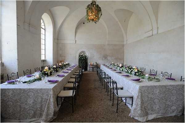 A wedding reception tablescape setup inside a vaulted stone hall with arched ceilings and whitewashed walls, photographed as a wide interior shot with no guests present. Two long rectangular banquet tables are dressed in white damask-patterned linen tablecloths and set with purple napkins on white plates, flanked by black wrought-iron cross-back chairs. Low floral centerpieces featuring cream and yellow flowers with trailing greenery run along the center of each table, accompanied by white taper candles. A chandelier draped in greenery hangs from the central vault, and an arched alcove at the far end of the room is decorated with a lush greenery installation. The decor palette combines purple, white, and green against the pale stone interior. Potential venue feature image.