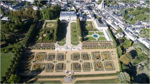 Aerial wide shot of a large French chateau and its formal gardens, showing a symmetrically designed parterre layout with geometric box-hedged beds, gravel pathways, a central lawn, and a circular pool. The white classical chateau building sits at the far end of the grounds, bordered by the surrounding town. Two small groups of people are visible on the garden pathways, likely guests or a wedding party, though they are very small relative to the scale of the grounds. The formal French garden design features precisely clipped hedgerows arranged in repeating square and rectangular patterns. Potential venue feature image.