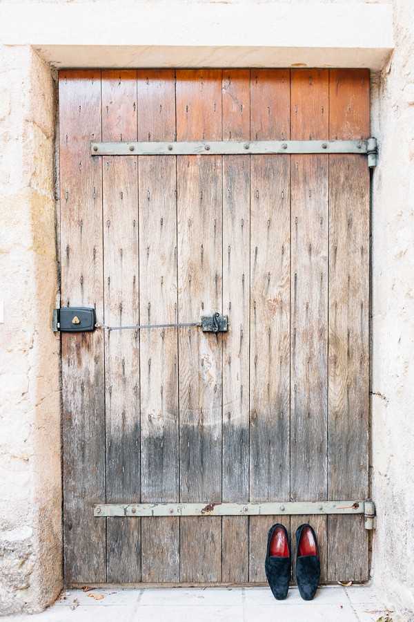 A getting-ready detail shot showing a pair of black velvet loafers with red insoles placed at the base of a large, weathered wooden door with metal strap hinges and a black iron latch mechanism. The door is set within a pale stone surround, suggesting a historic French property. The composition is a centered wide shot with the shoes as the focal point at the bottom of the frame, styled against the rustic backdrop as a groom's accessory detail.