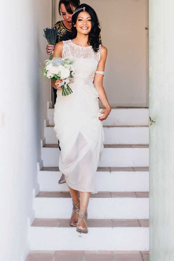A bride descends a white staircase accompanied by a woman behind her, likely a family member or bridesmaid, in a getting-ready or pre-ceremony moment. The bride wears a tea-length ivory dress with a sheer lace illusion neckline, a draped chiffon skirt, strappy gold heeled sandals, a pearl or crystal bracelet cuff, and a gold beaded headpiece. She carries a compact bouquet of white roses, succulents, and soft greenery. The second woman wears a black and gold sequined top and holds what appears to be a small bundle of dried lavender. The setting is an interior stairwell with white painted walls and tiled steps, consistent with a Mediterranean or South of France style property. The overall styling has a 1920s-inspired, art deco aesthetic. Portrait shot with natural light.