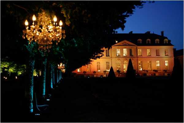 A nighttime exterior shot of a French château captured at dusk, with the building illuminated by warm amber uplighting that highlights its classic 18th-century architecture, symmetrical windows glowing from interior light, and manicured topiary cones visible along the façade. In the foreground, a tree-lined allée is decorated with tall candelabra-style crystal chandeliers mounted on stands, casting warm golden light along the pathway. The wide-angle composition frames the chandelier-lit avenue leading toward the château in the background. Potential venue feature image.