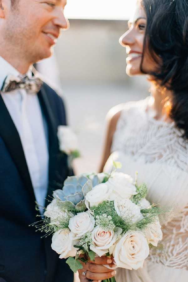 A close-up portrait of a bride and groom looking at each other and smiling outdoors in warm golden light. The groom wears a navy suit with a patterned floral bow tie, while the bride wears a lace dress with fringe or feather detailing along the neckline. The bride holds a bouquet composed of cream and blush roses, white ranunculus, succulents, baby's breath, and eucalyptus foliage. The composition is tightly cropped, keeping both faces partially in frame while centering the bouquet in the lower portion of the image.