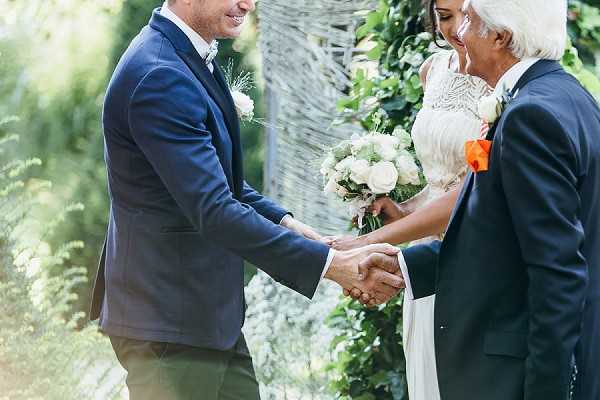 An outdoor ceremony moment in which the groom, wearing a navy blue suit with a white boutonniere, shakes hands with an older man in a dark suit who has a bright orange boutonniere, likely the bride's father during a hand-off or greeting. The bride stands between them in a white lace dress, holding a bouquet of white roses and eucalyptus. The backdrop features a woven willow or twig arch decorated with green foliage. The shot is a medium close-up focused on the handshake, with all three figures partially cropped.
