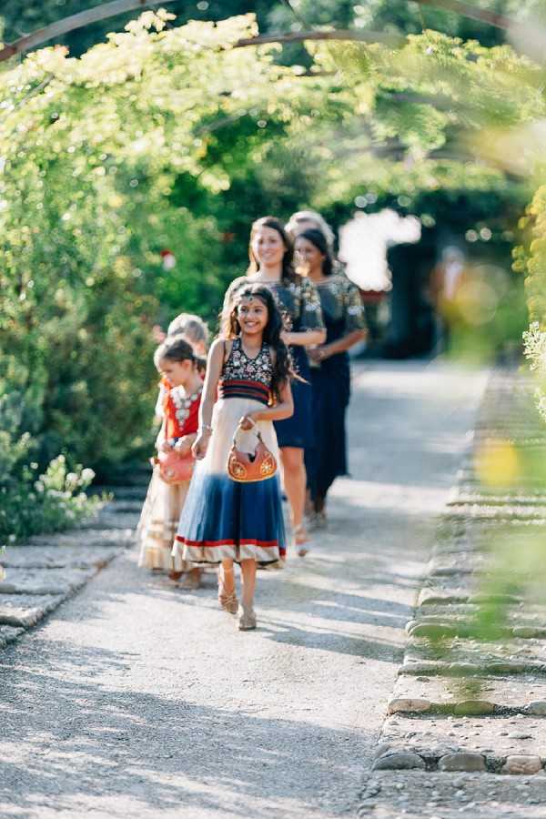A processional shot taken outdoors showing a group of approximately five people — including two young girls and two or three adult women — walking along a gravel path beneath a curved green pergola arch covered in foliage. The girl in the foreground wears a navy, cream, and red embroidered lehenga with a decorative maang tikka headpiece and carries a small ornate handbag, while the younger girl behind her wears a red and gold traditional Indian outfit. The adult women in the background wear navy floor-length dresses with embellished tops. The styling reflects an Indian-fusion wedding aesthetic with traditional South Asian dress elements. This is a medium wide shot with a shallow depth of field, keeping the foreground child sharp while the background figures are softly blurred.