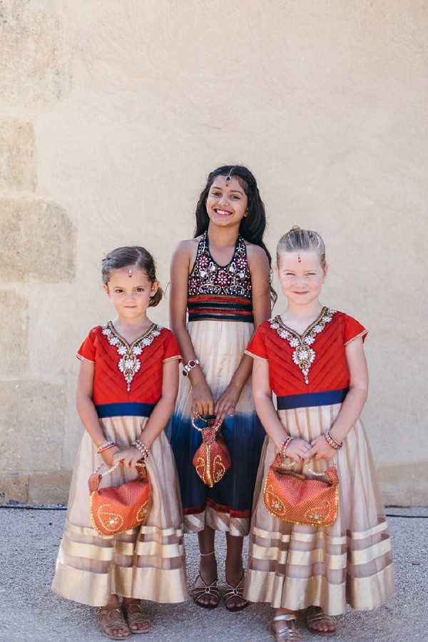 Three young girls pose together for a portrait against a pale stone wall, dressed in traditional Indian-style wedding attire. The center girl wears a navy and cream embroidered halter-style lehenga with a maang tikka head piece, while the two younger girls on either side wear matching red and gold lehengas with jeweled neckline embellishments, gold-striped cream skirts, and small orange beaded handbags. All three girls wear red bindis on their foreheads and gold bracelets, coordinating with a South Asian wedding aesthetic. This is a close-up portrait shot with all three children facing the camera.
