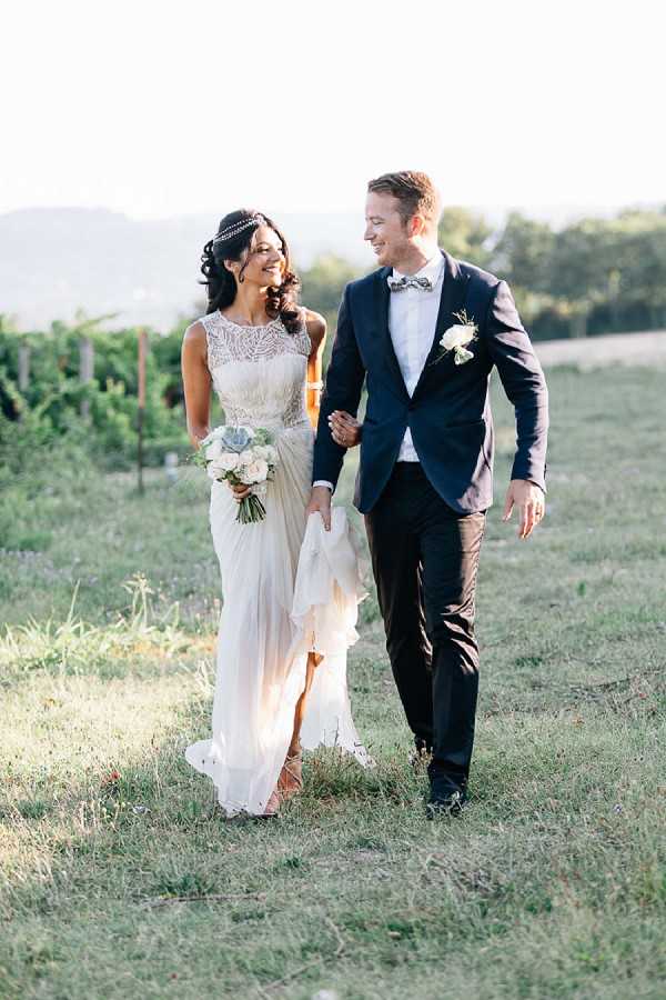 A couple portrait taken outdoors in a vineyard setting during golden hour light. The bride wears a sleeveless ivory gown with a lace illusion neckline and flowing chiffon skirt, accessorized with a beaded headpiece, and carries a bouquet of white roses, succulents, and greenery. The groom wears a navy blazer with black trousers, a patterned bow tie, and a white rose boutonniere. The two are walking and laughing together, captured in a full-length portrait shot with vineyard rows visible in the soft-focus background.