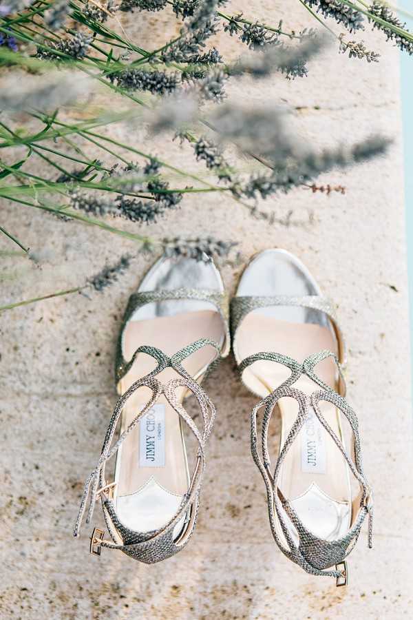 A detail close-up flat lay of a pair of Jimmy Choo bridal heels with silver and gold glitter multi-strap ankle-tie design, placed on a stone surface with sprigs of lavender resting above them. The shoes feature multiple strappy crisscross straps, gold buckle closures at the ankles, and a nude leather insole with the Jimmy Choo London label visible inside both shoes. The composition is shot from directly above, with soft natural lighting creating gentle shadows from the lavender stems across the stone surface.