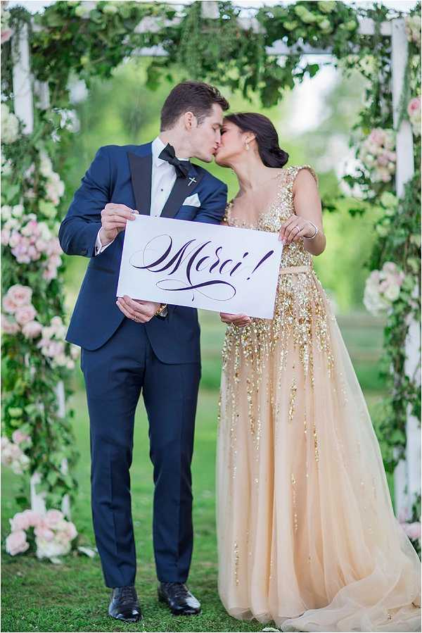 A couple kisses while jointly holding a white calligraphy sign reading 'Merci!' in an outdoor garden setting. The groom wears a navy blue tuxedo with black lapels, a white dress shirt, and a black bow tie, while the bride wears a champagne and gold heavily sequined ball gown with a deep V-neckline and cap sleeves. They are standing in front of a white pergola or arbor structure decorated with lush greenery and clusters of blush pink roses. The portrait is a medium full-length shot with soft natural light and a blurred green background.