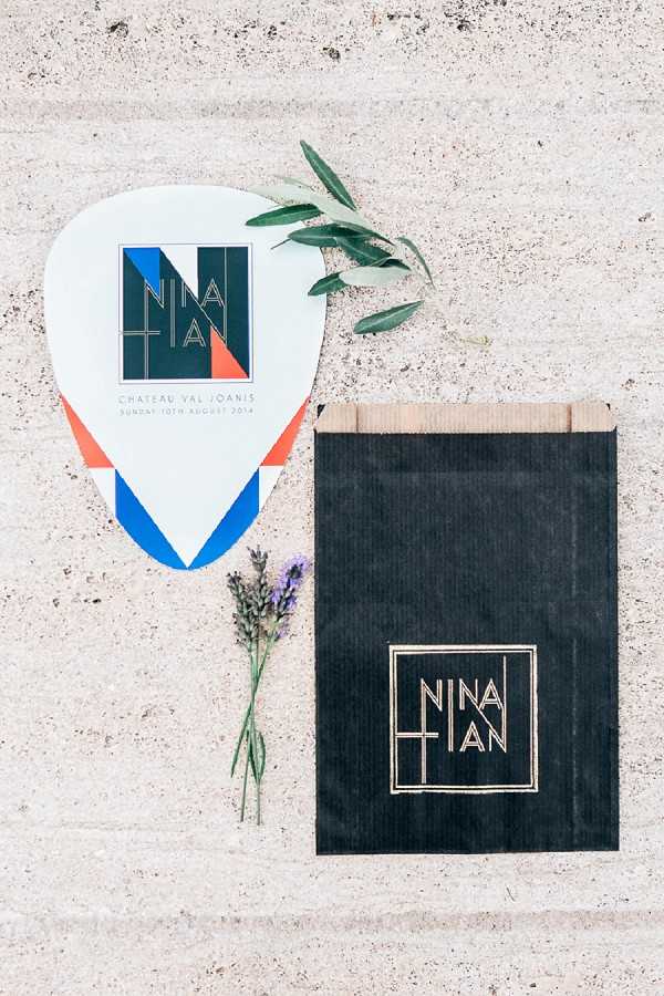 A flat lay detail shot of wedding stationery and paper goods arranged on a stone surface. The items include a guitar pick-shaped program or fan card in white, coral/orange, and cobalt blue featuring a geometric monogram logo for 'Nina + Ian' at Chateau Val Joanis, dated Sunday 10th August 2014, alongside a matte black paper bag with a gold foil 'Nina + Ian' monogram in a square geometric frame. A small sprig of lavender and an olive branch are placed between the items as styling accents. The overall design aesthetic is modern and graphic, with a bold geometric brand identity using a teal, coral, and blue color palette on the program contrasted with gold-on-black on the bag.