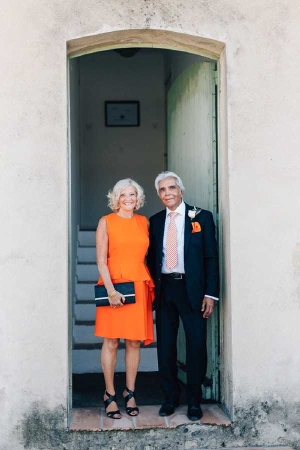 A portrait of two wedding guests — a woman and a man — posing together in an arched doorway of what appears to be a historic stone building, likely a French chateau or chapel. The woman wears a bright orange sleeveless peplum dress with black strappy heels and carries a black clutch, while the man wears a dark navy suit with a red-and-white striped tie, an orange pocket square, and a white boutonniere accented with an orange flower, coordinating their outfits. The doorway frames a glimpse of interior stone stairs and a green door behind them. The shot is a full-length portrait with a vertical composition.