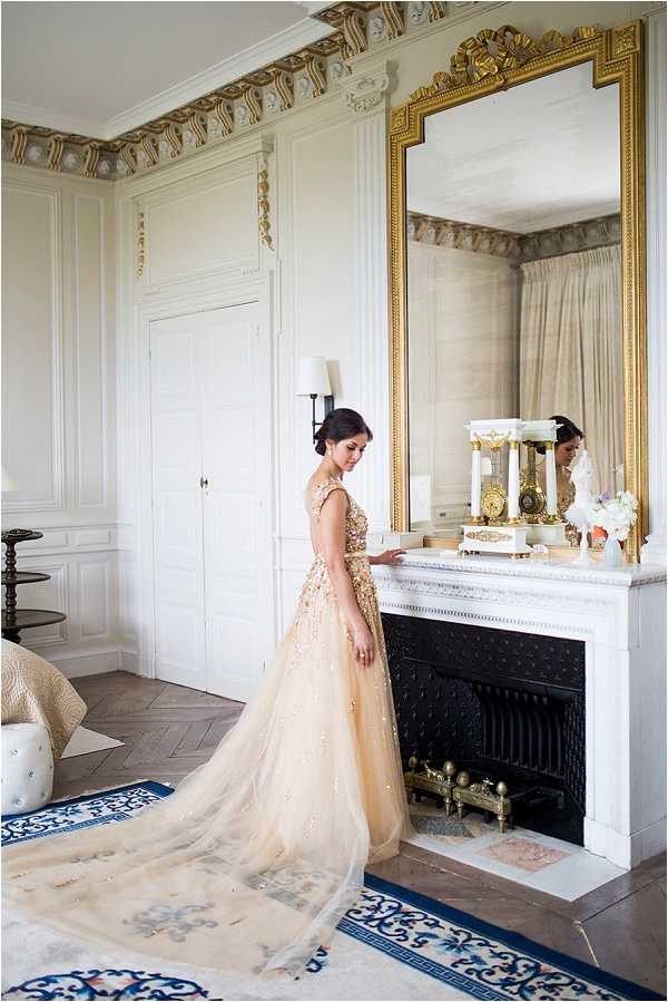 A bride stands alone in an ornate interior room during what appears to be a getting-ready or portrait moment, resting her hand on a white marble fireplace mantel. She wears a champagne-gold ballgown with heavily embellished floral appliqué on the bodice and a long, flowing tulle skirt with a full train. Her dark hair is styled in an updo. The room features white-painted paneled walls with gilded decorative molding along the cornice, herringbone parquet flooring, and a blue-and-white patterned area rug. A large ornate gold-framed mirror above the mantel reflects the bride from a different angle, also revealing she wears a veil. A white marble mantel clock with column detailing and gold candlestick accents sit on the mantelpiece. The overall interior style is classic French château or Haussmann-era apartment décor. The shot is a full-length portrait taken from the side.