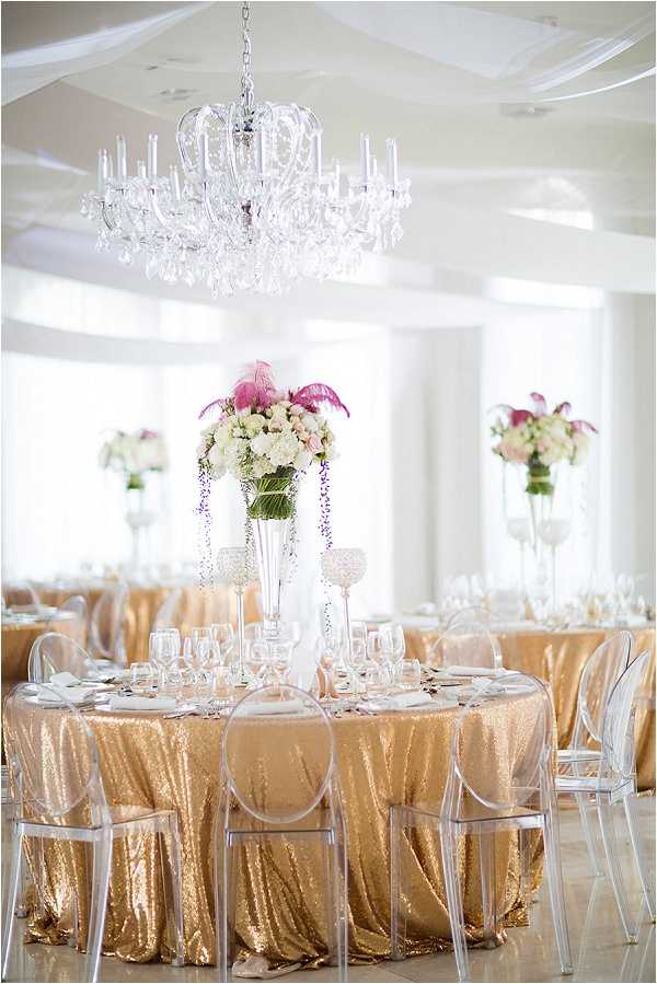 A wide shot of a wedding reception ballroom setup with no guests present. The indoor venue features white walls, white ceiling draping, and a large crystal chandelier as the central overhead light fixture. Round tables are dressed in gold sequin floor-length tablecloths and surrounded by clear acrylic ghost chairs. Tall trumpet-vase centerpieces hold arrangements of white hydrangeas, blush roses, trailing purple wisteria, and hot pink feathers. Additional crystal candleholders and clear glassware are placed on each table setting. Multiple similarly styled tables are visible in the background, creating a consistent gold, white, and pink decor palette throughout the room. The overall styling is formal and classic with a maximalist approach to floral height and table linens.