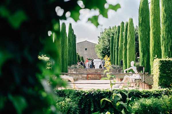 A wide shot captured through soft foreground foliage showing a cocktail hour or pre-ceremony gathering at a formal garden venue in the South of France. A group of approximately 10–15 guests in colorful attire are visible in the middle distance near stone steps leading up to a Provençal stone building. The garden features tall, narrow cypress trees arranged in formal rows alongside neatly clipped low hedgerows, creating a structured, classical French garden layout. Two white sculptural figures are positioned on a low stone wall in the right foreground, suggesting intentional contemporary art installations as part of the venue's decor. Potential venue feature image.