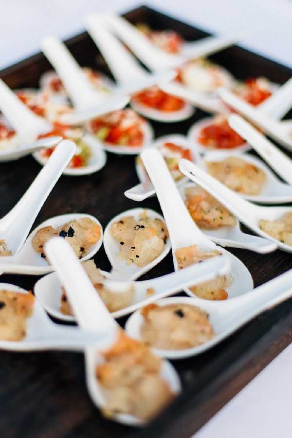 Close-up detail shot of cocktail hour canapés arranged on a dark wooden serving tray. Two varieties of bite-sized appetizers are presented on white ceramic tasting spoons: the foreground rows hold a warm-toned seafood or poultry preparation with black seasoning, while the background rows hold a red tomato-based topping, likely a bruschetta-style bite. The spoons are arranged in neat diagonal rows across the tray, creating a repeating pattern. The shallow depth of field keeps the foreground spoons in sharp focus while the background blurs progressively.
