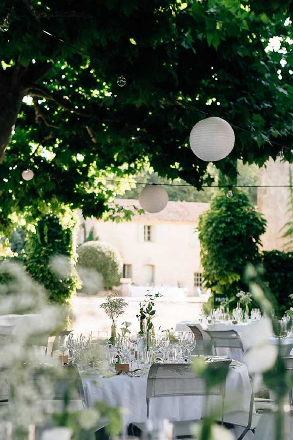 An outdoor wedding reception setup photographed from a medium distance, with round tables covered in white or light grey linens arranged beneath a canopy of dense tree foliage. Table centerpieces consist of tall, slender glass bud vases holding delicate white florals and green foliage, likely baby's breath and eucalyptus, with multiple wine glasses and place settings visible. White paper globe lanterns hang from string lights strung between the trees, and a warm-toned stone Provençal farmhouse building is visible in the background through the greenery. The styling is minimal and natural, with an all-white and green palette, consistent with a rustic-chic or garden-party aesthetic. Potential venue feature image.