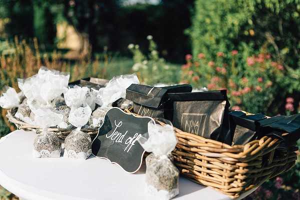 A close-up detail shot of a wedding send-off table set outdoors, with a garden visible in the background. A small white table holds a wicker basket filled with dark-packaged branded goods labeled 'Nina' alongside individually wrapped favors in white translucent tissue, likely containing dried herbs or lavender, tied with small bundles. A black chalkboard-style sign with gold script reads 'Send off,' indicating these items are intended for guests departing the event. The styling uses a mix of natural textures (wicker, dried botanicals) and dark packaging for a rustic-modern aesthetic.