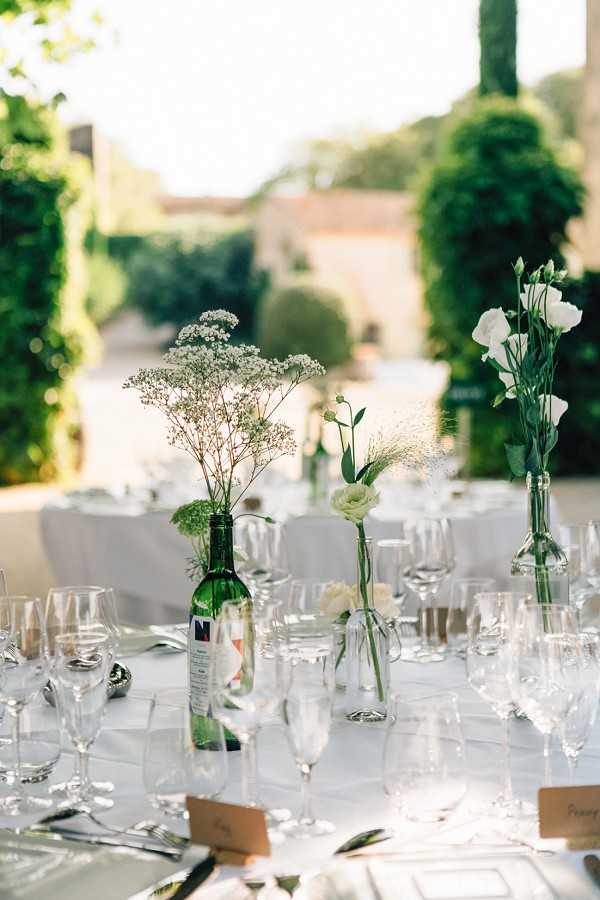 Close-up detail shot of an outdoor wedding reception table set with white linen, crystal glassware, and silver cutlery. The centerpiece styling uses repurposed green wine bottles and clear glass bud vases holding a mix of white Queen Anne's lace, white lisianthus, a cream ranunculus, and wispy dried grasses, creating a casual, garden-inspired aesthetic in a white and green palette. Small kraft paper place cards are visible at the table settings. The setting appears to be a French property with manicured topiary and hedging visible in the soft-focus background, suggesting an outdoor terrace or courtyard reception.