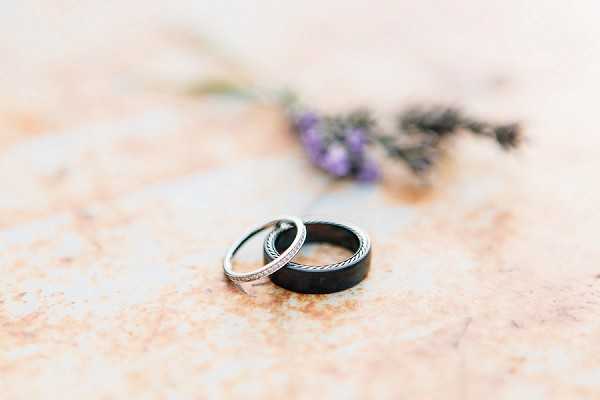 A close-up detail shot of two wedding bands resting together on a weathered, rust-toned surface. One ring is a slim silver band with pavé diamond detailing, and the other is a wide dark gunmetal or black band with a rope-twist textured edge. In the soft-focus background, a small sprig of dried lavender and a pale blue ribbon are visible, adding a Provençal styling touch to the flat-lay composition.